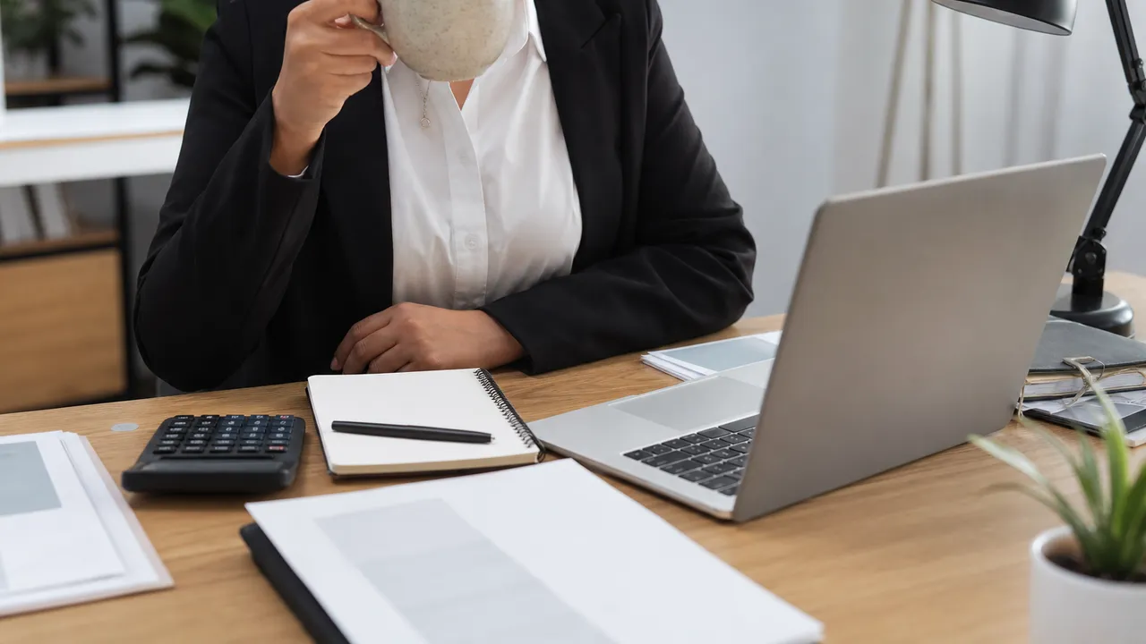 Personne en tenue de travail prenant un café devant son ordinateur portable, carnet et calculatrice posés sur le bureau